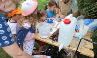 Children gather around a table filled with craft materials. In the background, an adult works with clay as the children look on with curiosity.