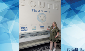 Member of staff in a green dress standing in front of a case labelled 'South: The Antarctic' which contains fossils and rocks. 