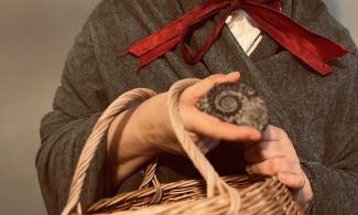 An actress playing the palaeontologist Mary Anning. She is wearing a shawl and the red ribbon of her bonnet is visible, her face is not in the picture. She is holding a basket and an ammonite. 
