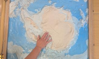A 3D map of Antarctica in a wooden frame, showing the contours of the mountain ranges and surrounding islands. A hand is reaching out to touch the ridge of the Transantarctic Mountains near the Ross Ice Shelf. 