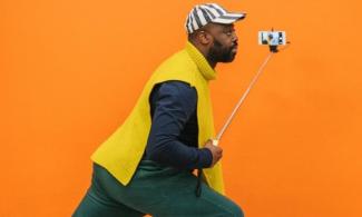 A man stands side-on, holding a selfie stick with a smartphone attached, facing toward the phone. He is wearing a bright yellow sleeveless jumper over a dark shirt and a black-and-white striped cap. The background is a solid, vivid orange.