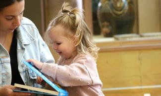 alt= "a young girl in a pink jumper opens a story book with her mum on her right"