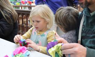 A little girl with her tongue slightly poking out in concentration sits at a table surrounded by other children and adults as she creates a flower using tissue paper.