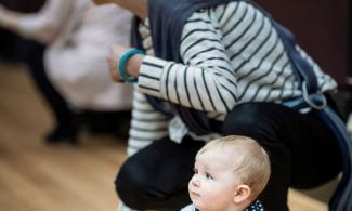 alt='a baby sat on the floor in a blue shirt, his mother crouches behind"