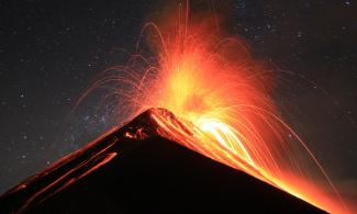 An erupting, cone-shaped volcano, its lava glowing orange against a starry night sky