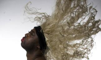 Harold Offeh wearing a long curly blonde wig, bouncing in the wind behind his head.