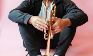 A man dressed in black sat in a pink room, holding a trumpet.