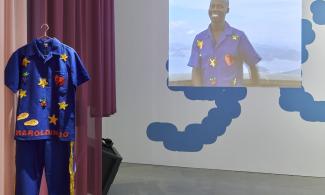 A video project of Harold Offeh wearing a royal blue boiler suit, decorated with patches. The costume hangs near the video projection.