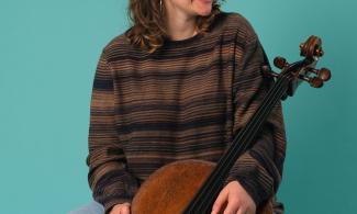 A woman with shoulder length brown hair, dressed in a brown striped jumper, holding a cello and smiling.