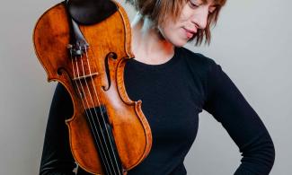 A woman white a brown bob haircut looking down at the floor. In her right hand she holds a violin upside down.