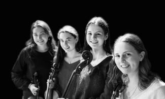Four women standing in a line, smiling. They are each holding a string instrument.