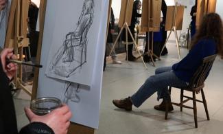 alt="a model with long dark brown hair dressed in blue sits on a wooden chair to the right of the image, seven people stand around her on the left of the image painting her on their easels" 