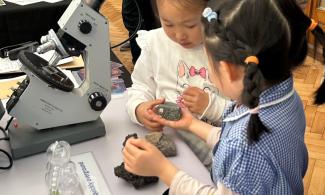 Two young girls look carefully at a rock together, with one of them holding it in the palm of her hand. They are standing by a table with a microscope on it, and other rocks