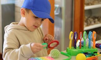 A young boy in a blue hat and beige hoody holds a magnifying glass up to a fidget toy. Other fidget and sensory toys lay on the table in front of him. 