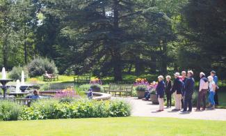 A group of people stands near a circular fountain in a lush garden filled with colorful flowers and greenery. Benches are scattered around the area, with large trees in the background under a clear, sunny sky.