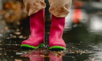 A person wearing bright pink rain boots with green soles stands in a puddle on a wet street. Fallen leaves are scattered around, and there are reflections in the water.