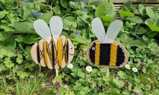 Two handcrafted bee decorations made from cardboard and yarn are displayed among green foliage and small wildflowers. The bee on the left has yellow and black yarn, while the one on the right has black and yellow stripes. Both have paper wings and googly eyes.