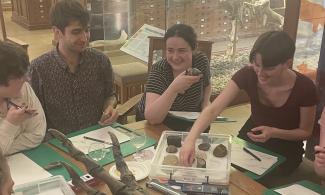 A group of adult visitors to the museum are smiling and chatting around a table. On the table is a range of sketching materials and a box of fossils. Visitors are holding fossils that they have picked out to sketch. A replica foot fossil of a allosaurus, a large carnivorous dinosaur, is also on the table.