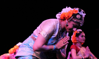 Dancers on stage in their costumes after performing an Indian classical dance. The performers have their hands pressed together as two of them bow to the audience.