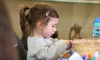 alt= "a young girl with long brown hair tied in a ponytail plays with arts and crafts supplies in a wicker basket"