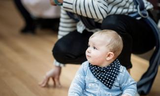alt='a baby is sat on the floor in a blue shirt and blue spotted necktie, his mother crouches behind"