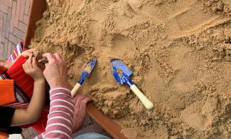 Image of a sand pit with a blue spade and the hands and arms of an adult and child finding pottery shards