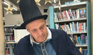 Image shows a man wearing a black top hat and jacket writing on a piece of piece or parchment with a quill.