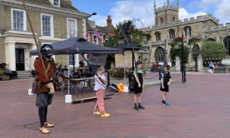 Image of three children following instructions from an adult dressed up as a Civil War soldier on a market square, with a church in the background