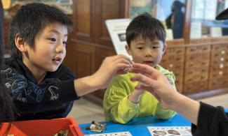 A child is holding out a fossil they have found to a Sedgwick Museum volunteer for identification help. Their younger sibling looks on in the background.