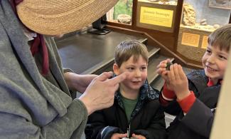 To children visiting the museum are smiling at an actor playing Mary Anning. One of them is holding up a fossil that Mary has just 'sold' to them.