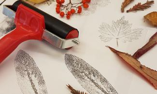 A table displays art supplies and nature-themed prints. There's a red roller, sheets with leaf prints and various dried leaves and twigs.