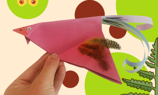A hand holds up a pink origami bird in flight, with realistic feathers added to the wings and curled paper tail feathers
