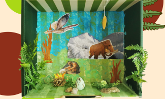 The interior of a large shoe box has been painted to form a scene with blue sky and green grass. To the right hand side is a snow covered mountanous area with a wooly mamouth. To the left is a stone age man lighting a 3D fire, adjacent is a  3D painted foam egg. The scene also has a prehistoric bird in flight and 3D foligae stuck to the sides of the box.