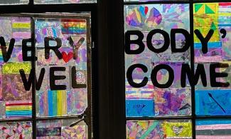 The words 'EVERYBODY'S WELCOME' are written across a colourful window, which is slightly open. The window is mad up of a rainbow of plastic coloured transparencies, created by visitors to the museum. The patters they form celebrate queer love and Pride, including the Pride flag. Close up, the transparencies are thin sections of rock, with the different mineral crystals within a rock sample being a different colour.
