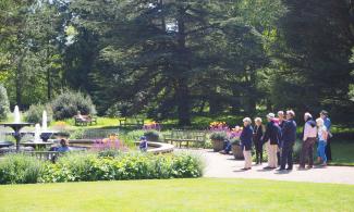 A group of people stand in the distance in a garden, looking at a fountain with various plantings and green lawn in front.