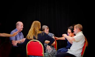 alt="a group of five adults sit in a circle facing each other on red chairs. They all have their hands in front of them or pointing out to the side. They are dancing sat down. The background is black"