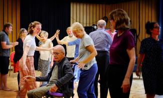 In the foreground a lady pushes a gentleman in grey with glasses and grey hair in a wheelchair. Behind them a group of ten people are dancing, facing various direction. Those facing the camera are smiling and enjoying themselves.