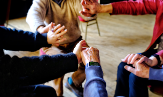There are four people sitting down facing inwards in a circle but you only see their hands, arms and two torsos. They are all clasping hands of the person next to them. 