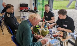 A photo of a table at a repair cafe. Four men are sitting round, smiling, repairing and writing things down. There is a bright window behind and a messy table of tools and papers.