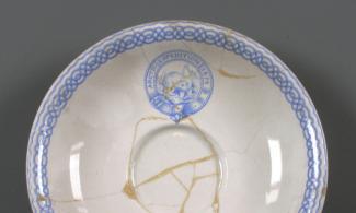 A detail view of one side of a cracked white dish with blue ornamentation around the rim. A blue circular crest is visible in the centre, with an image of a polar bear. It reads: 'Arctic Expedition 1875'. 