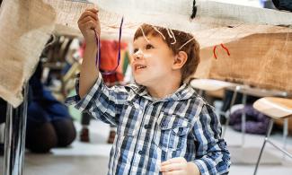 Small boy uder a linen parchment. He is playing with a thread, smiling to the upper left hand side of the screen.