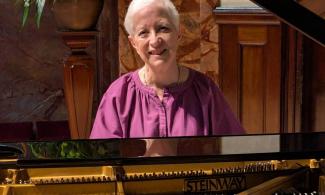 Susan Tomes wearing a purple blouse and smiling behind an open Steinway grand piano.
