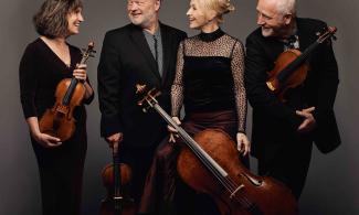 The four members of the Brodsky Quartet smiling at one another, holding their violins, viola and cello against a grey background.