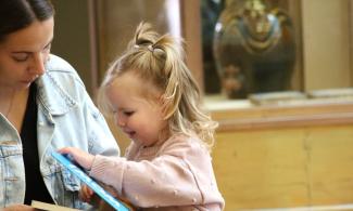 A baby, wearing pink, opens a book, with an adult with long hair looking on
