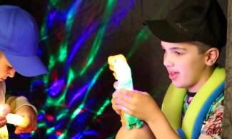 alt="two boys are sitting in a black tent, playing with light up dinosaur toys. The one on the left is dressed in a beige hoodie and blue cap and the one on the right is wearing a colourful t shirt with graffiti style writing on the arm, a black cap and wears a green and blue toy snake around his neck"