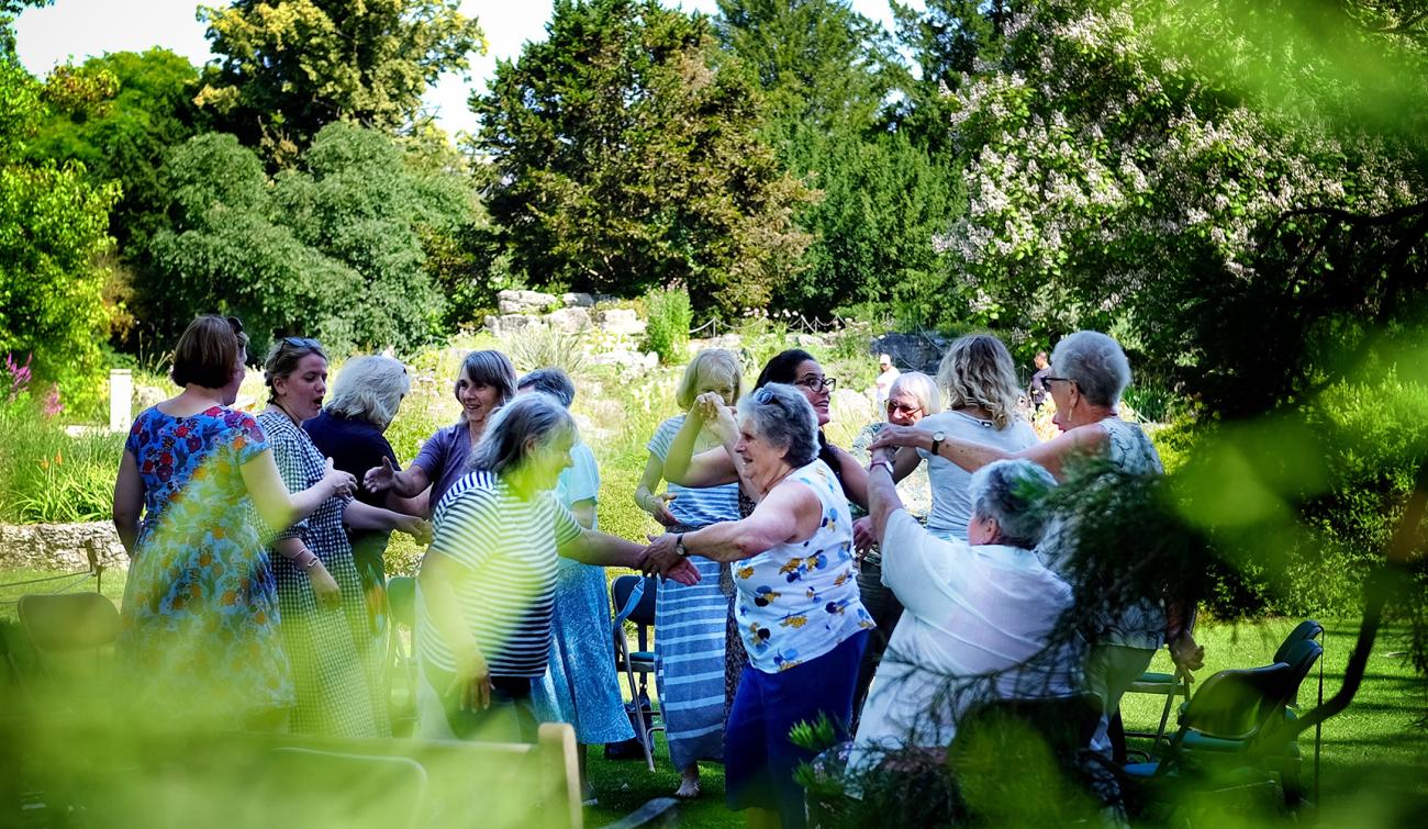 A group of adults stand smiling and dancing in a circle at the Cambridge University Botanic Garden.