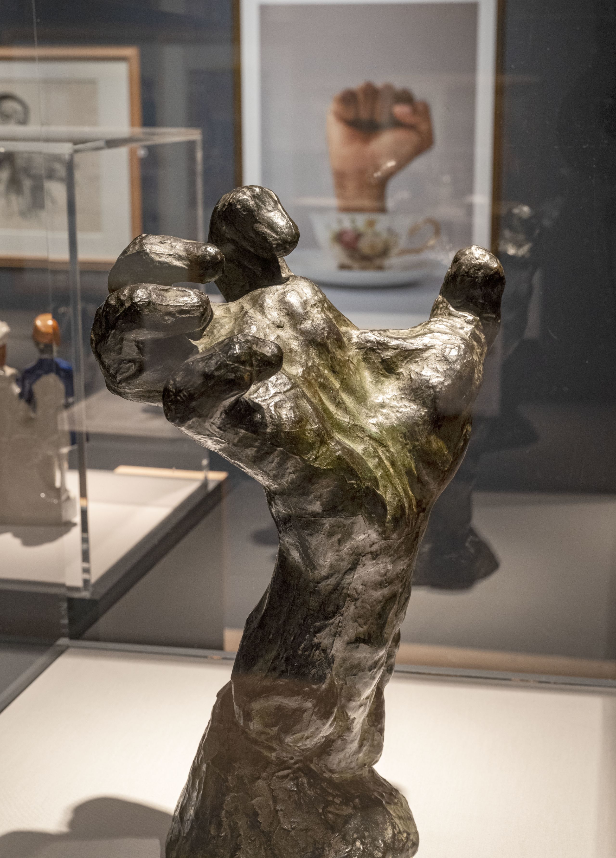 Bronze sculpture of a powerful right hand, with fingers contorted inwards. In the background is a large photograph of a fist punching up through a delicate tea cup