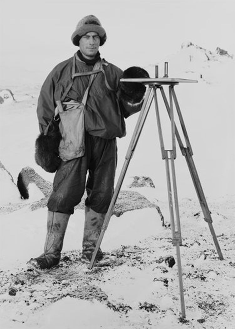 Frank Debenham, wearing sledging outfit, stands next to a plane table on a tripod.