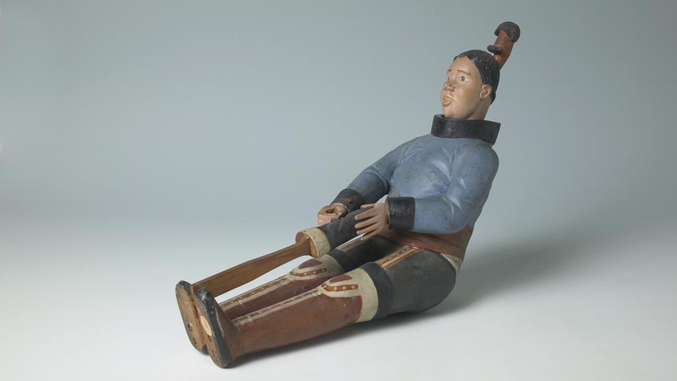 Carving of a woman, made of painted wood. She is sitting, leaning slightly backward and wearing a blue parka with black collar and cuffs and wide red waistband; black trousers with red and cream vertical stripes and cream dots on each side of the front; and thigh-length red boots with black soles and cuffs and cream decoration. Her black hair is tied in an upright knot painted black and red. Left hand is outstretched; right hand is clenched. She is holding the foot of a black boot with cream cuff. One end of a wooden stick is inserted into the boot; the opposite end is fixed between her feet.