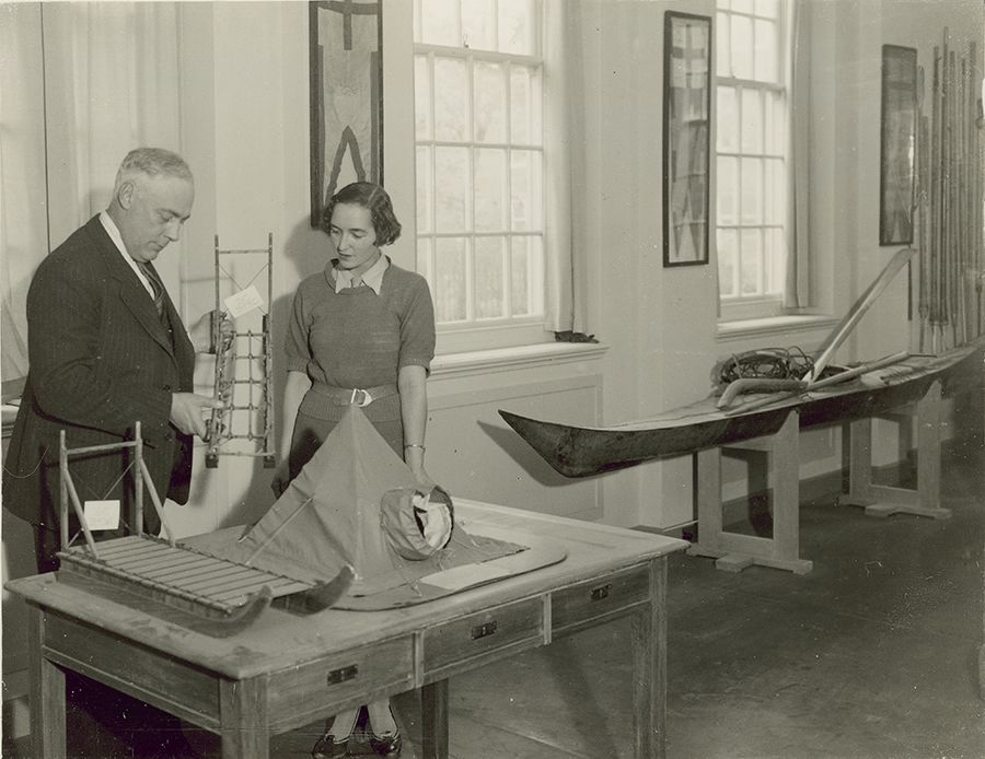 Frank Debenham and his assistant Elizabeth Creswick stand next to a table, on top of which is a model sledge and a model tent. To their right is a full size kayak.